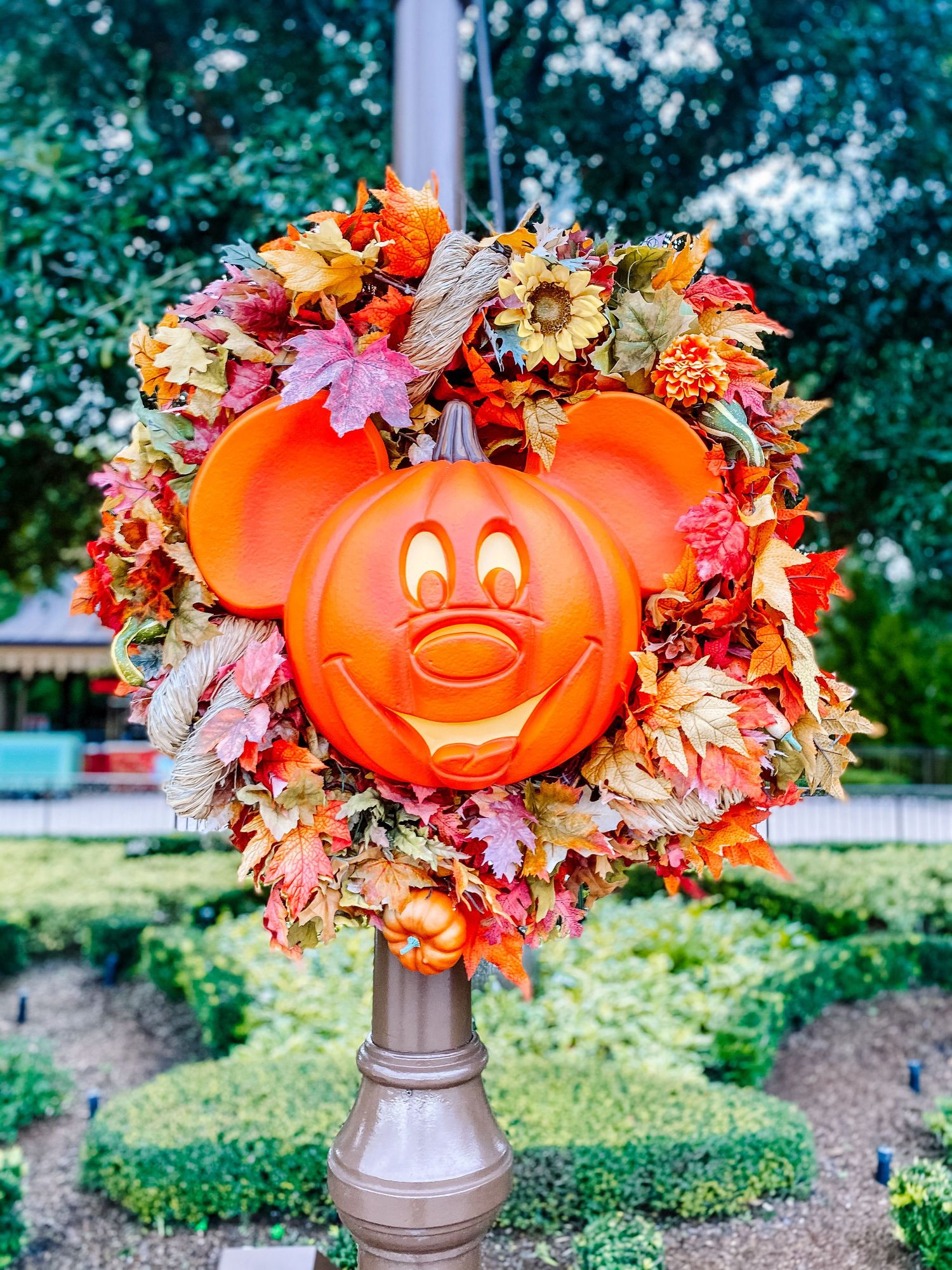 A festive Mickey Mouse pumpkin wreath with fall leaves and flowers adorns a pole in an outdoor garden setting.