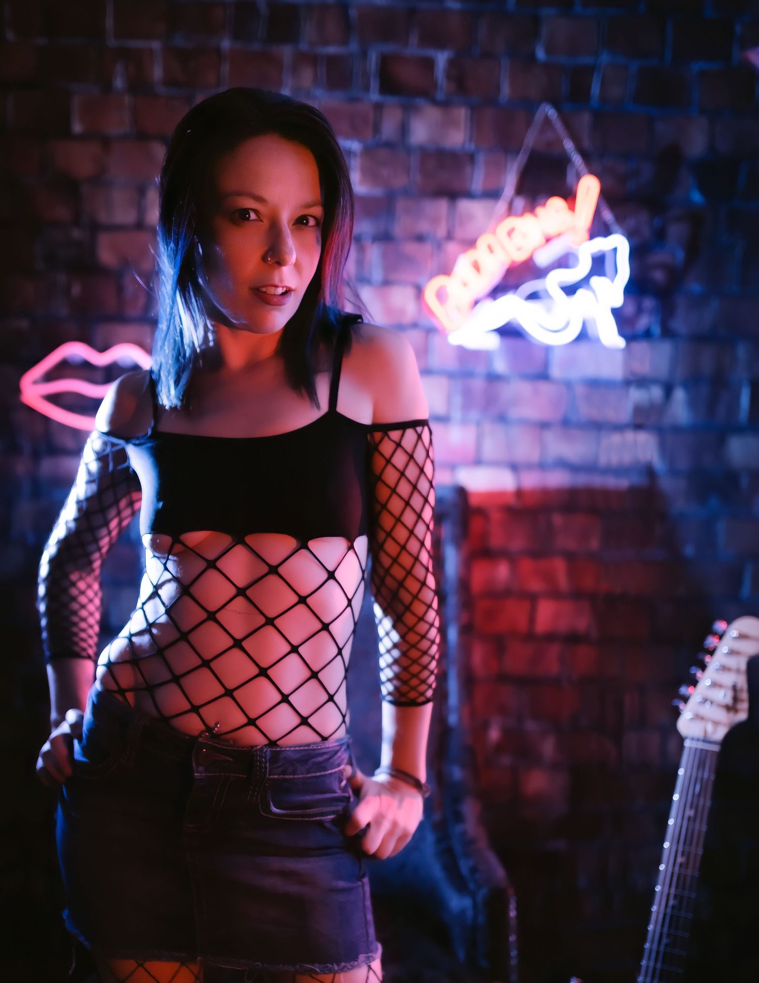 Female wearing a fishnet top with a jean skirt. Posing in front of a brick wall with neon signs and a guitar