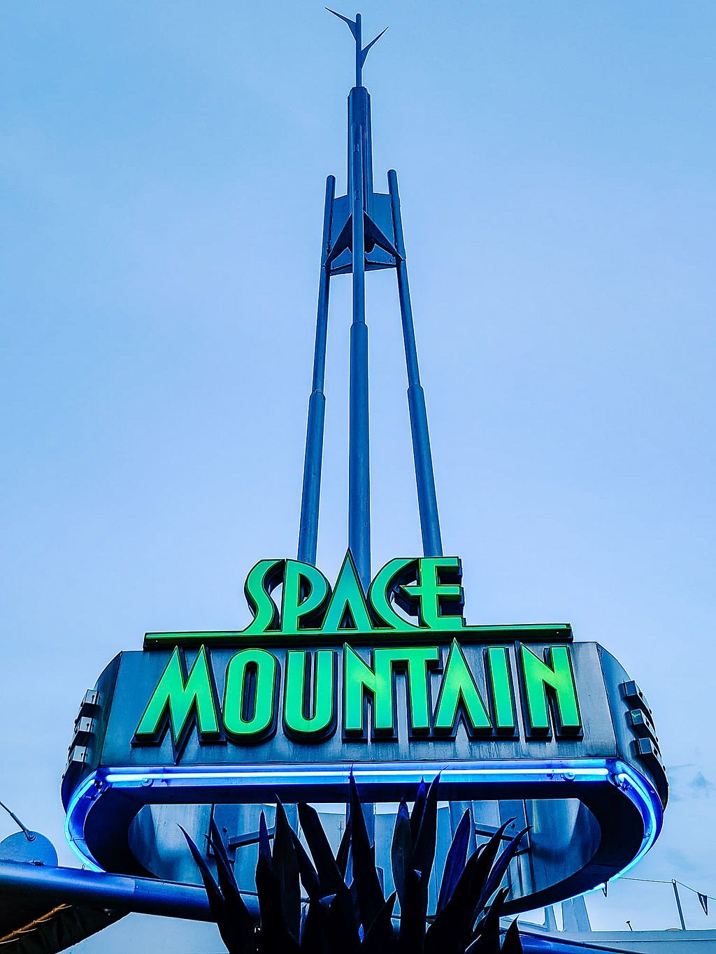 Iconic green neon Space Mountain sign glows against blue sky at Disneyland theme park.