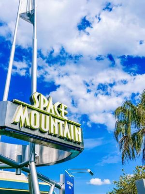 Space Mountain illuminated sign against bright blue sky with white clouds and palm tree at Disney theme park.