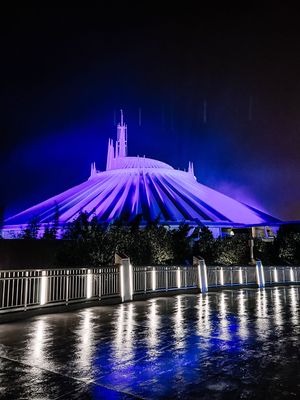 Illuminated Space Mountain ride at Disney World glows in purple and blue at night with reflections on wet pavement.