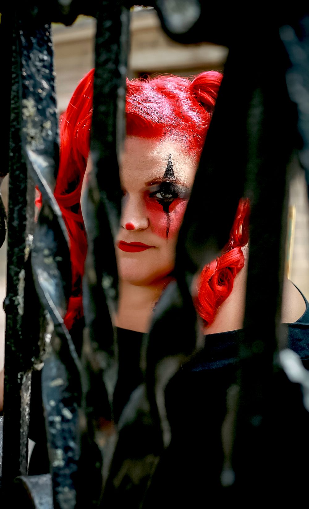 A dramatic Halloween costume with bright red hair and dark makeup is seen through iron bars in shadowy lighting.