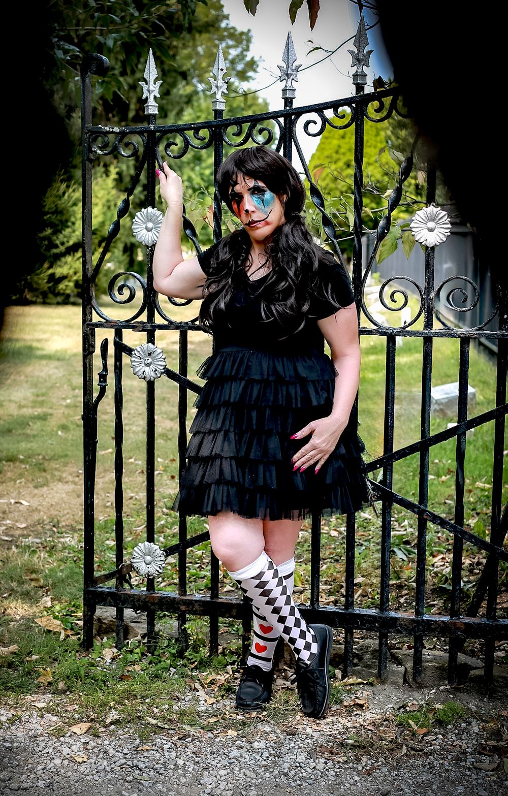 Gothic figure poses by ornate cemetery gate wearing black ruffled dress and striped knee socks.