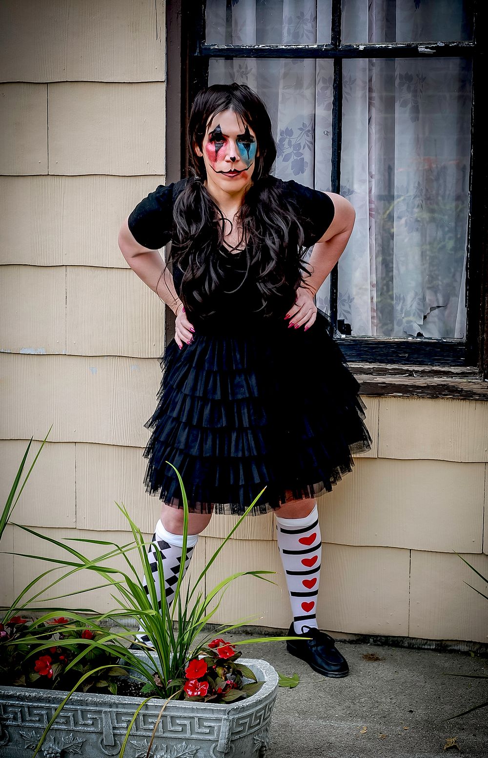 A figure in layered black dress and striped socks poses confidently by a potted plant.