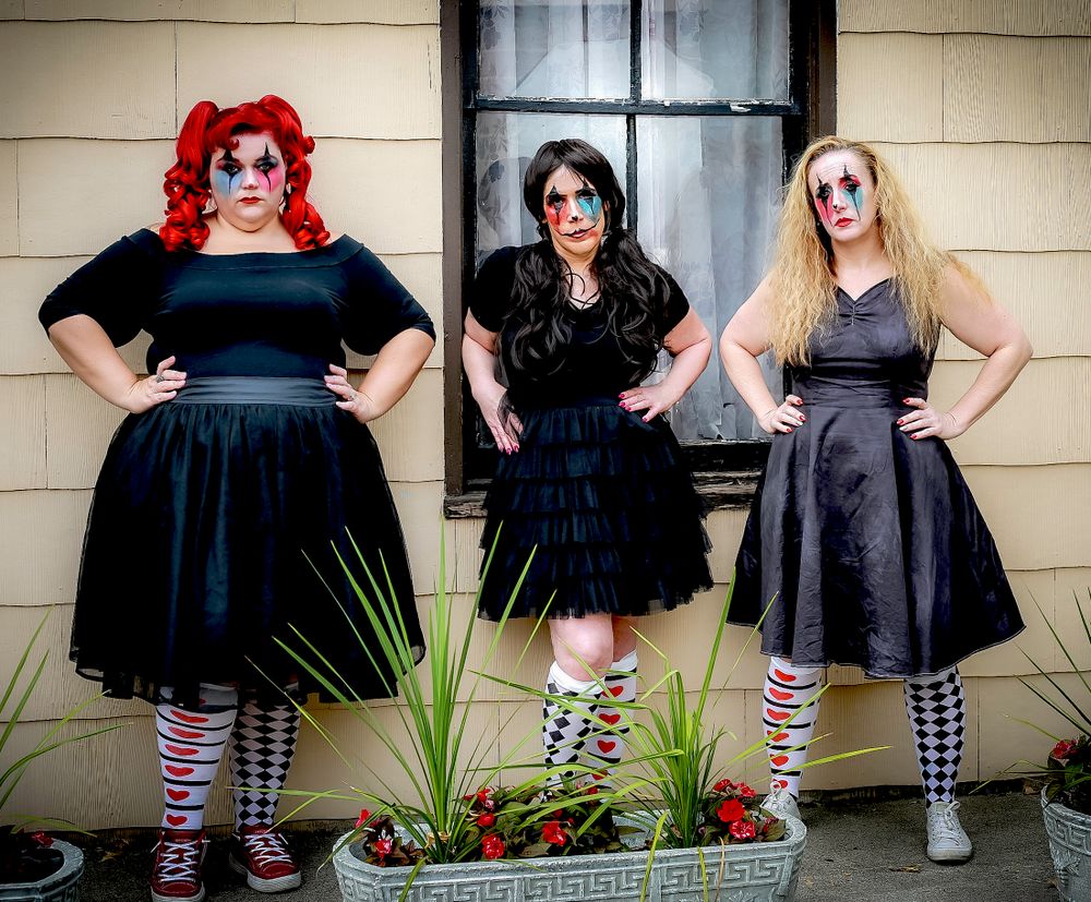 Three figures in gothic dresses and striped socks pose together in front of a weathered door.