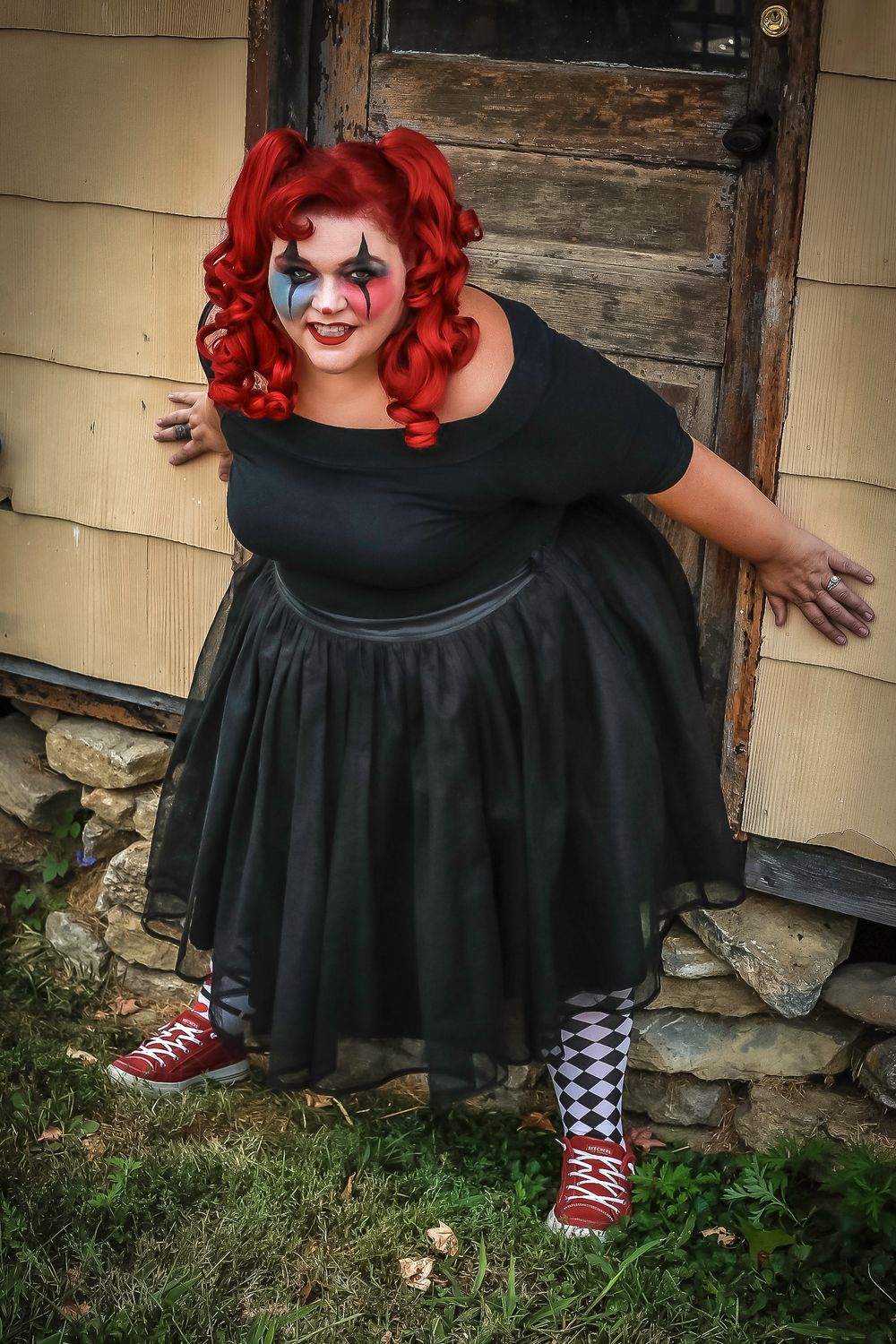 Figure in black dress and striped socks with bright red hair poses against rustic door.