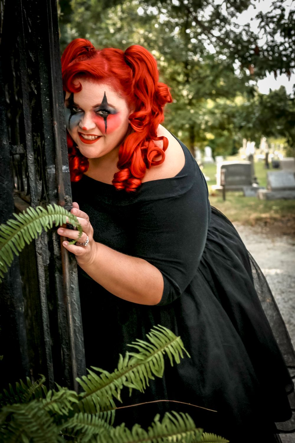A gothic figure with bright red hair poses playfully near greenery in a cemetery setting.