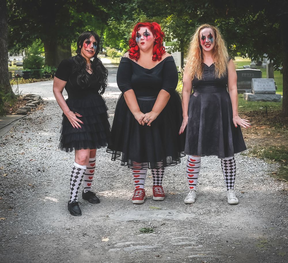 Three figures in dark dresses and striped socks stand together on a cemetery path.