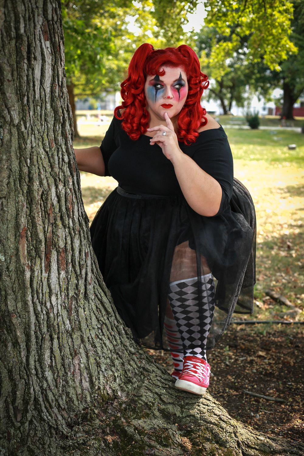 A gothic figure in black dress poses playfully against a large tree trunk.