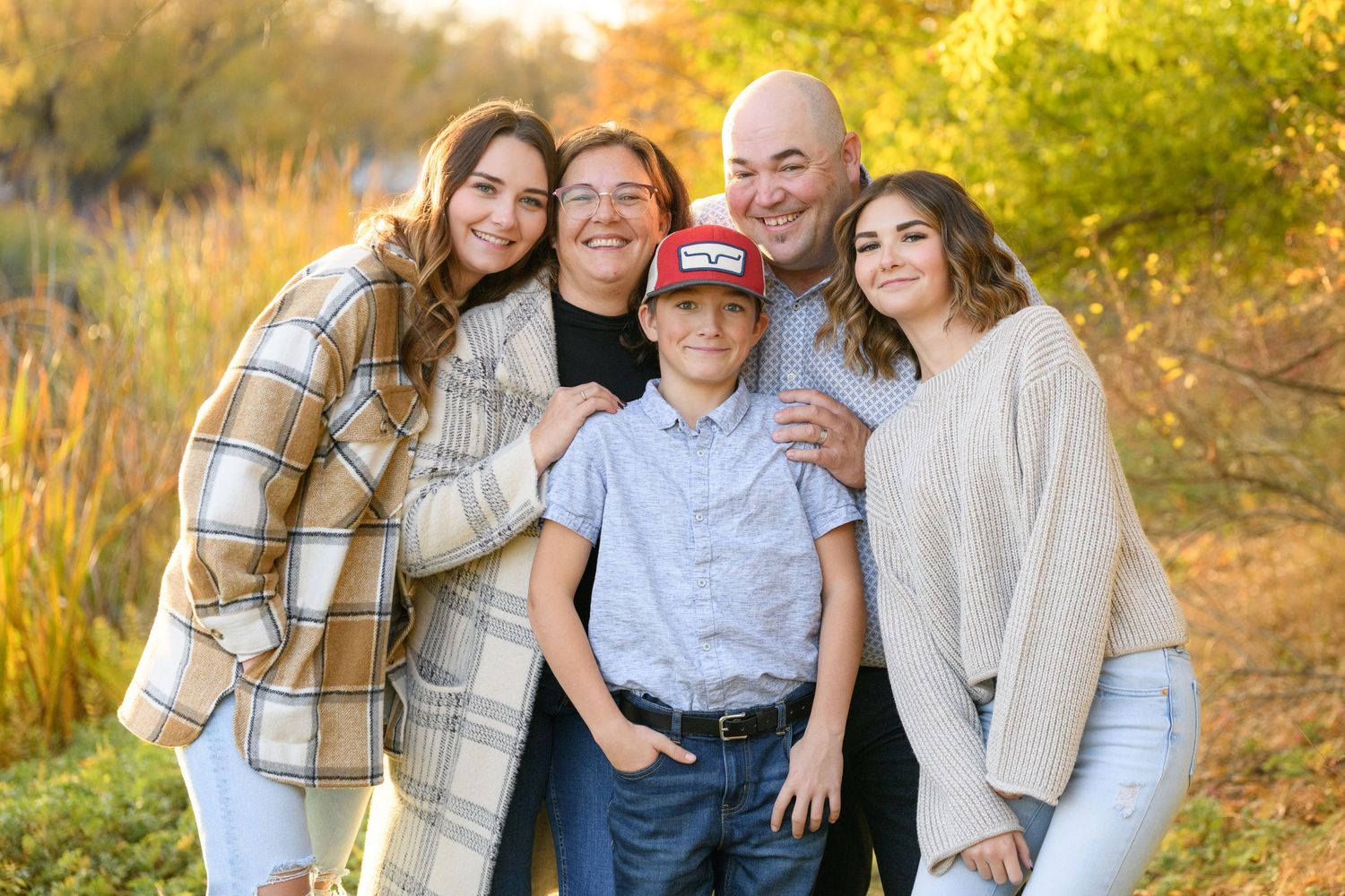Happy family in cozy sweaters poses together outdoors during golden hour autumn photoshoot.