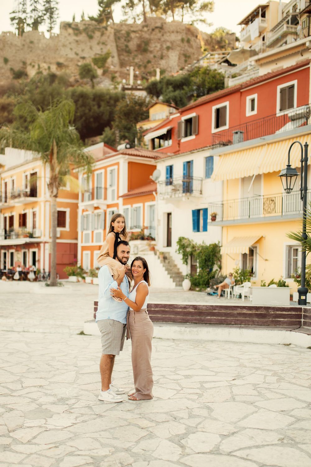 Three people embrace while standing on stone pavement with colorful coastal buildings behind them.