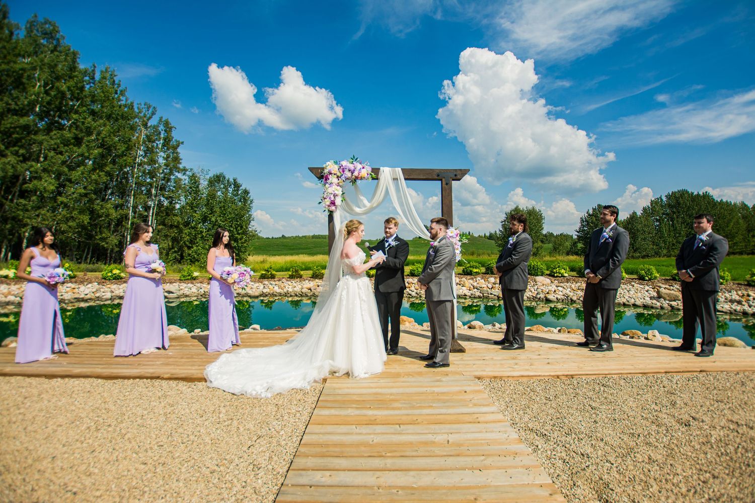 A couple in wedding attire embraces on a cliff overlooking a scenic mountain lake on a sunny afternoon. Garden View Estates Wedding Venue