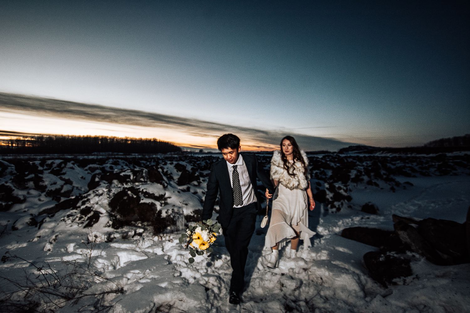 A couple in wedding attire embraces on a cliff overlooking a scenic mountain lake on a sunny afternoon.