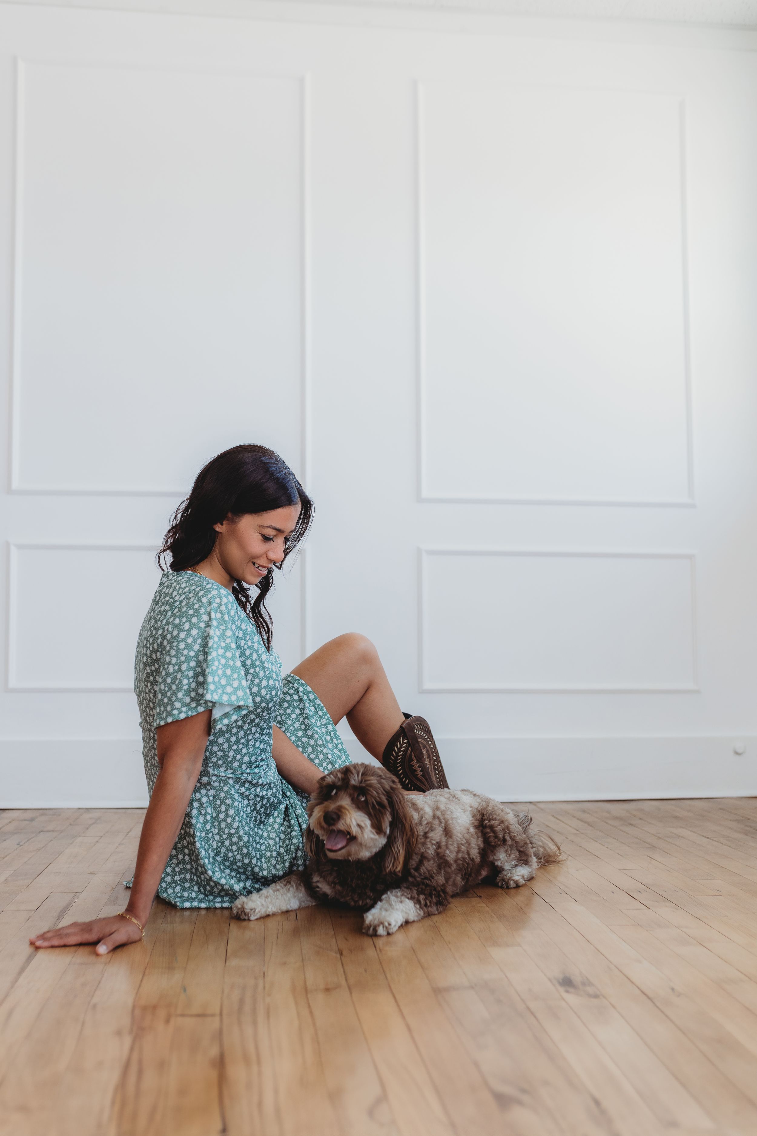 Woman in polka dot dress sits on hardwood floor with brown and white dog against white wall.