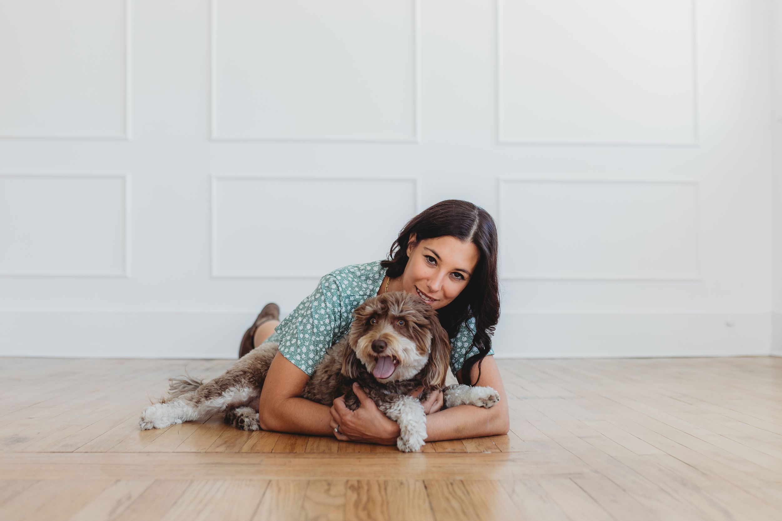 A happy brown and white dog lies on a hardwood floor with someone wearing a turquoise polka dot shirt.