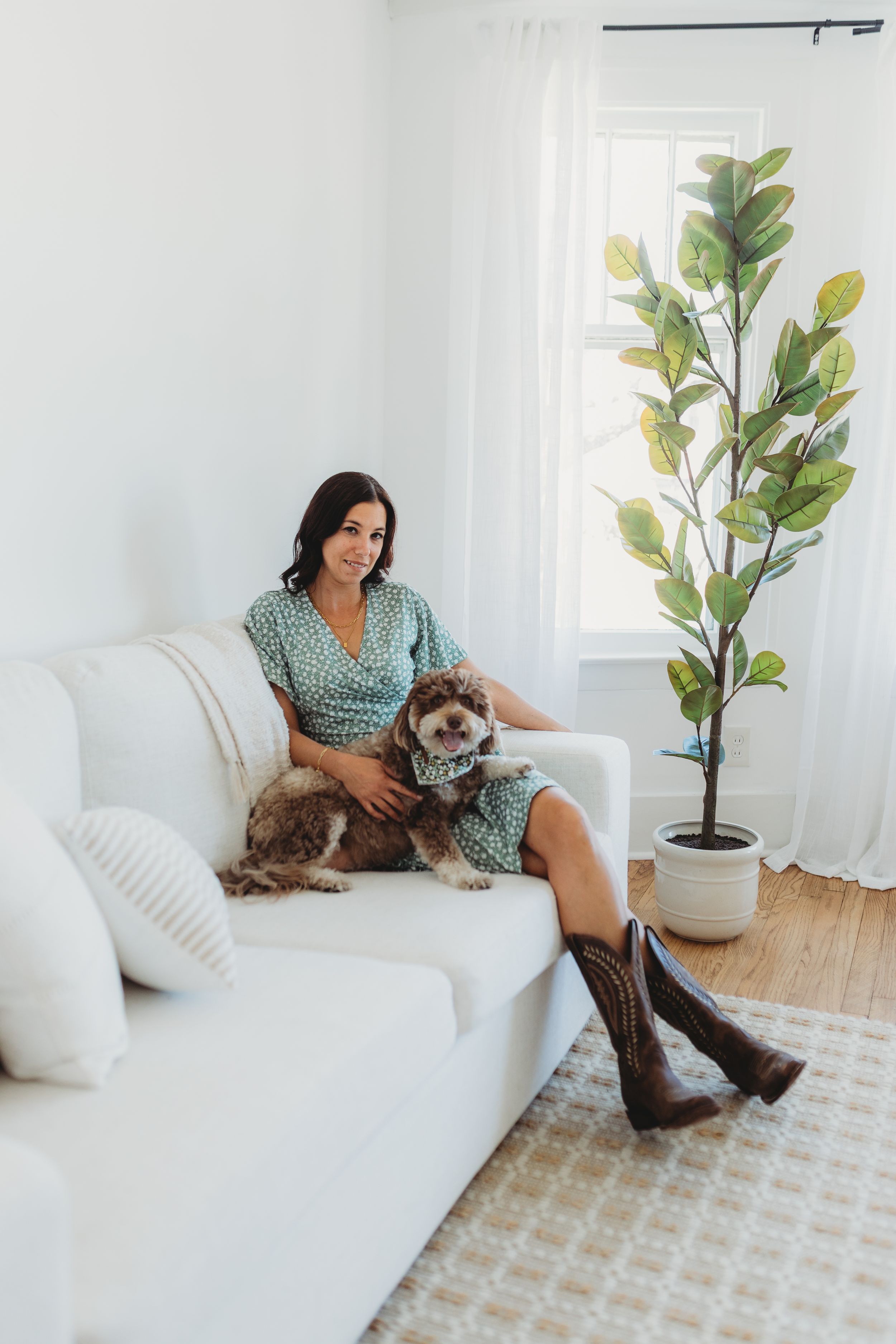 A cozy living room scene with a polka dot dress, brown boots, and a cute dog on a white couch beside a tall fiddle leaf plant.