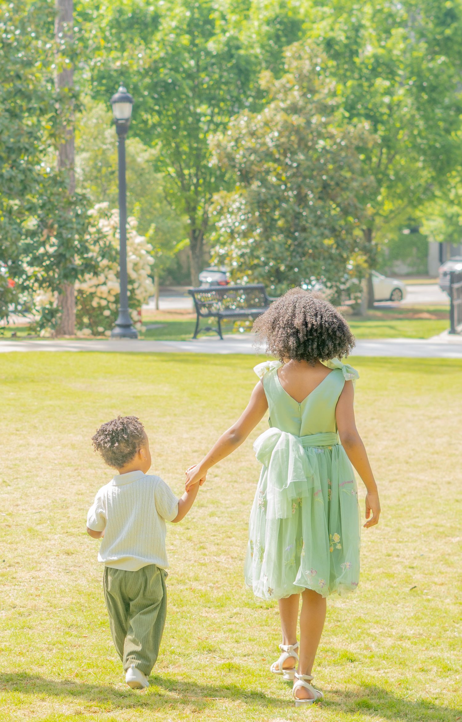 A parent and child hold hands while walking together through a sunny park with green grass and trees.