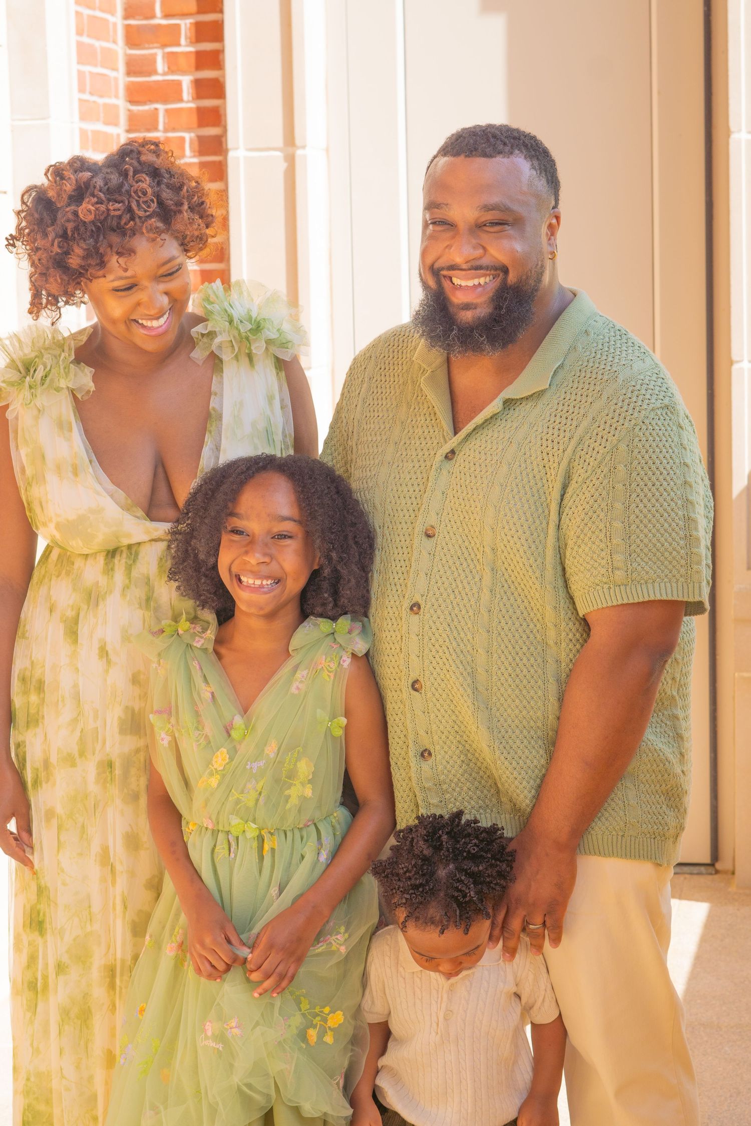 A family of four wearing coordinating yellow and green outfits poses together outdoors against a brick wall.