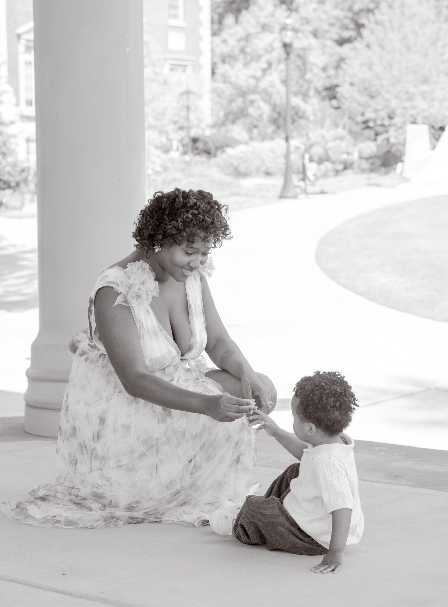 A tender moment captured in black and white as a figure in a floral dress sits with a small child on a porch.