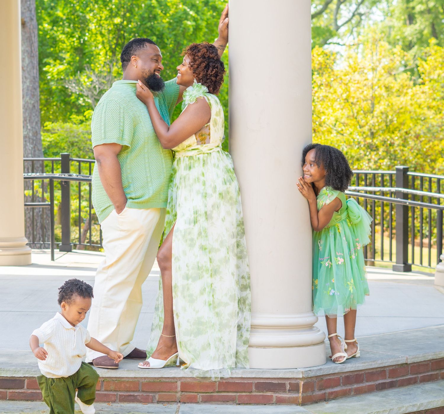 A family in coordinating mint green and white outfits poses together on a columned porch during a photo shoot.