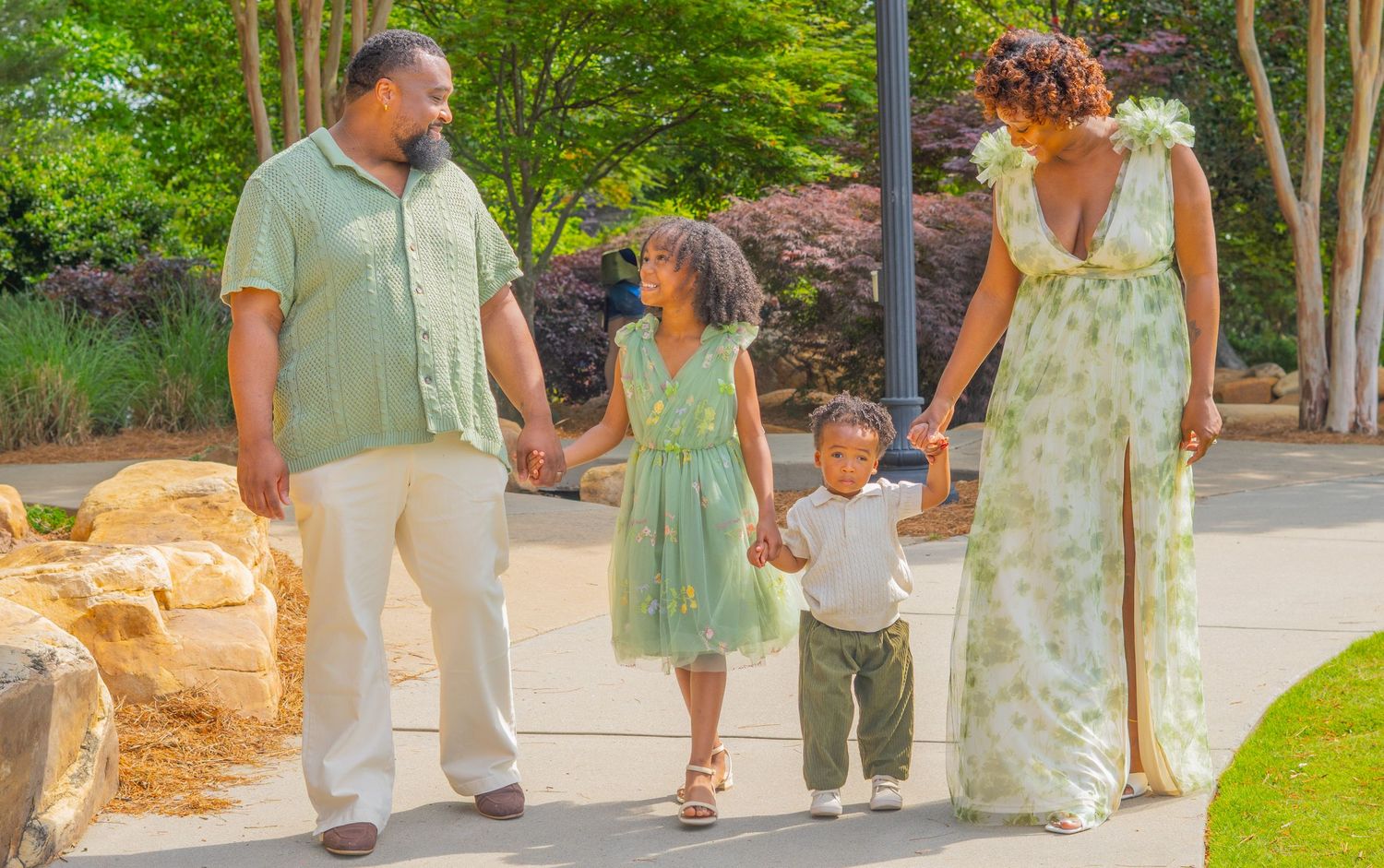 A family of four walks together on a garden path in coordinated summer outfits.