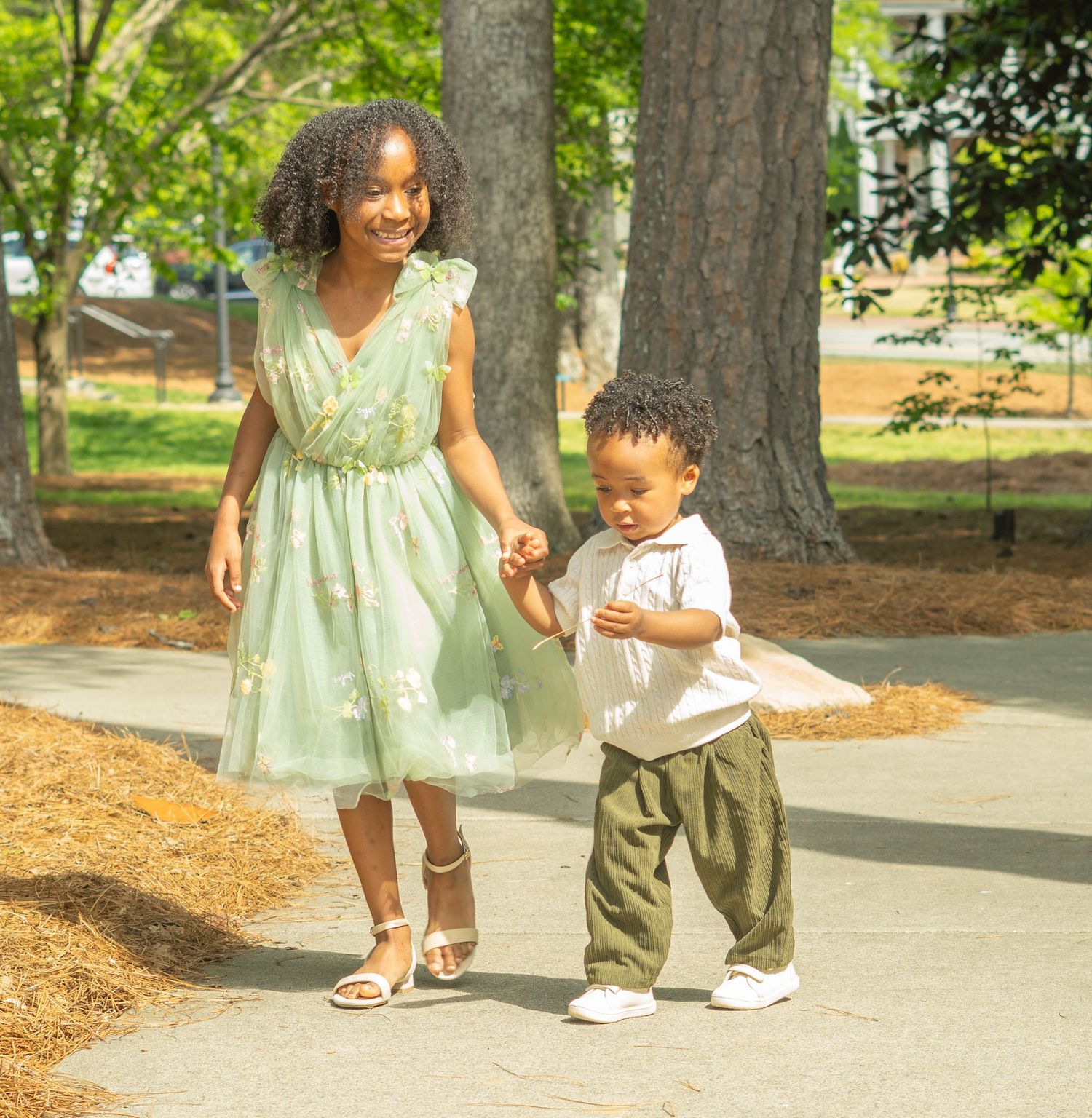 A woman in a mint green dress walks hand in hand with a toddler along a tree-lined path on a sunny day.