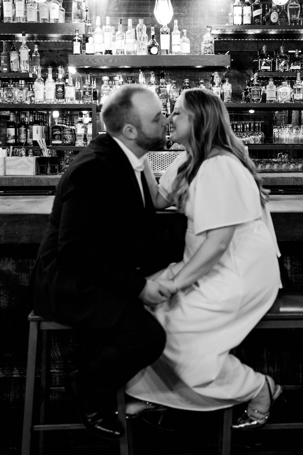 A romantic black and white photo of a couple sharing an intimate moment at a bar with bottles illuminated in the background.