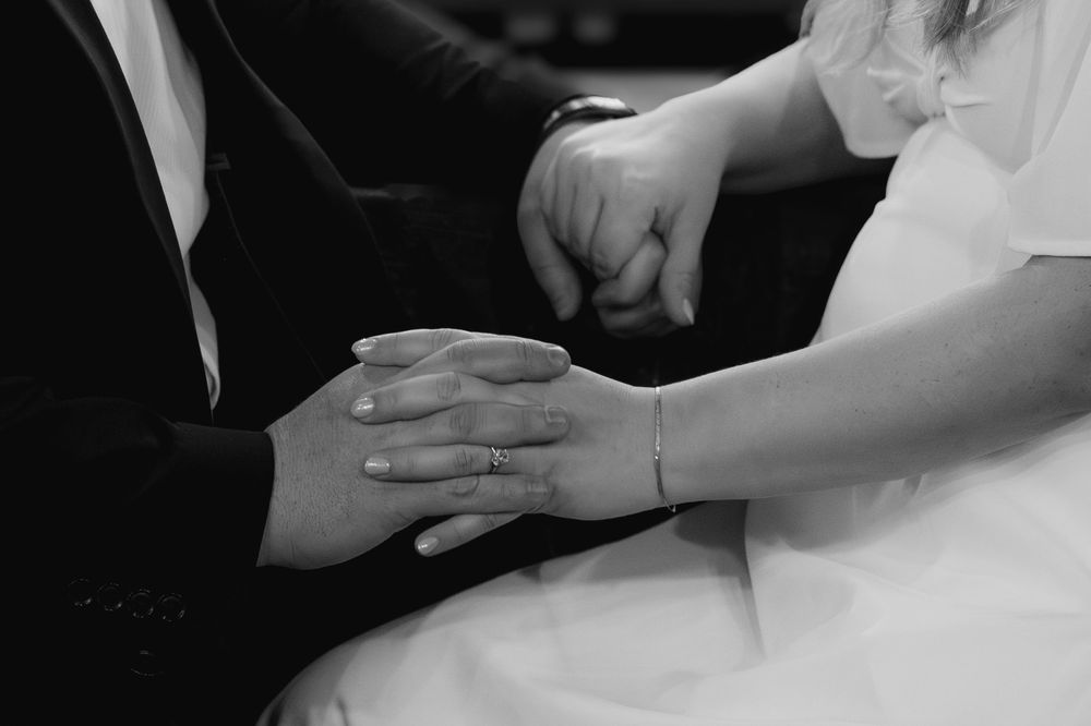 A black and white close-up photograph shows hands tenderly clasped together during a wedding ceremony.