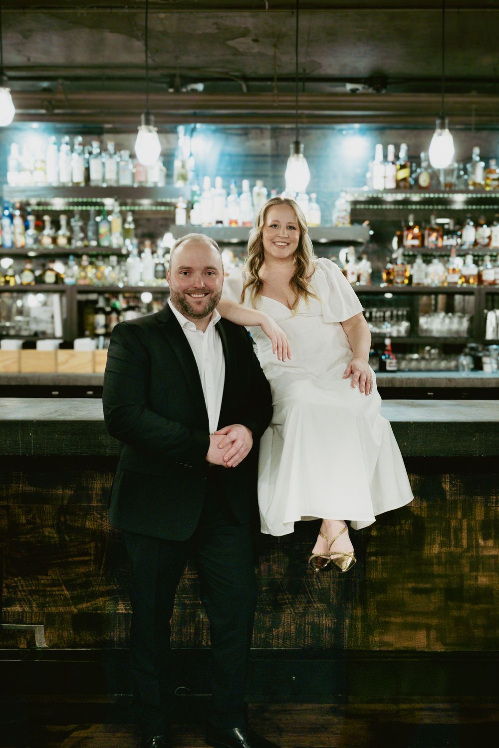 Couple poses at an elegant bar with dark wood and hanging pendant lights during a wedding celebration.