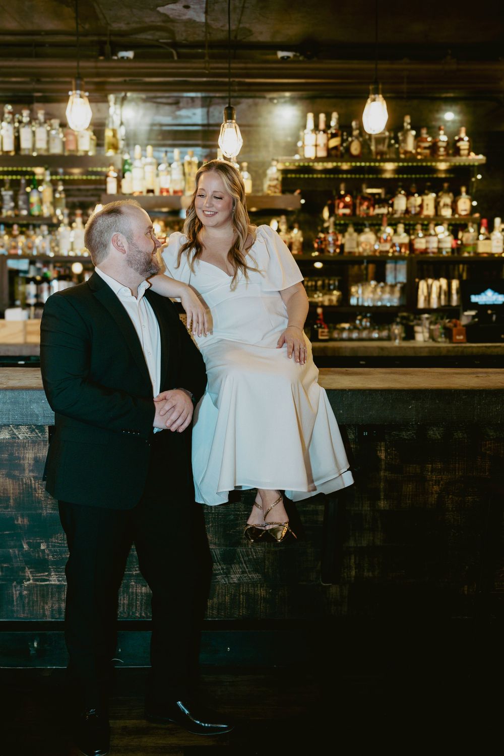 A wedding couple share a romantic moment at an elegant bar with illuminated shelves of bottles behind them.