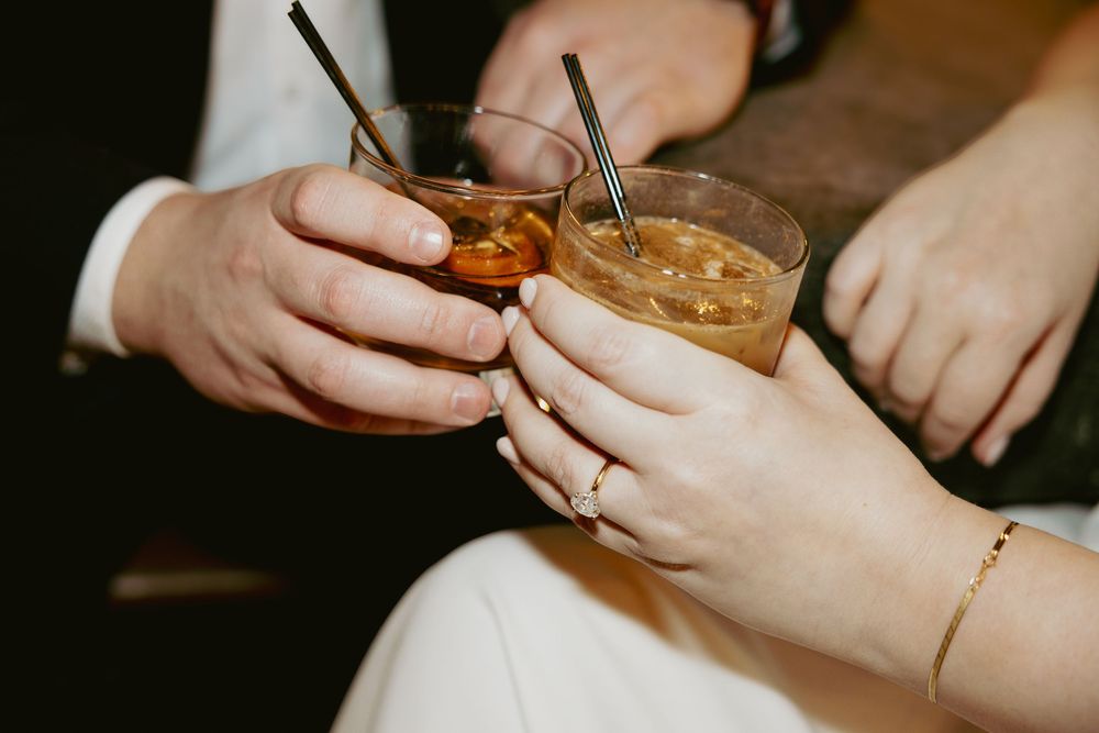 Two people clink glasses filled with amber-colored cocktails in a dimly lit setting.