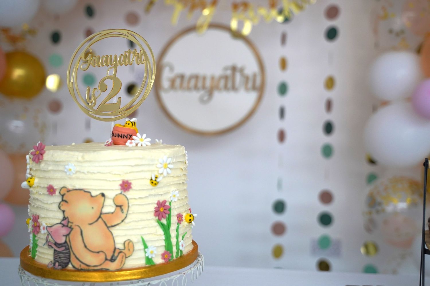 A decorated birthday cake with Winnie the Pooh design and gold cake topper displayed against a bokeh background.