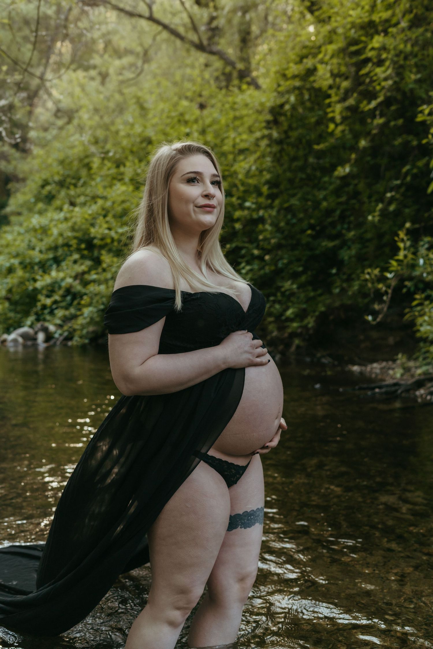 A maternity photo shoot by a creek shows an expectant mother in a black dress kneeling on the shore in natural lighting.