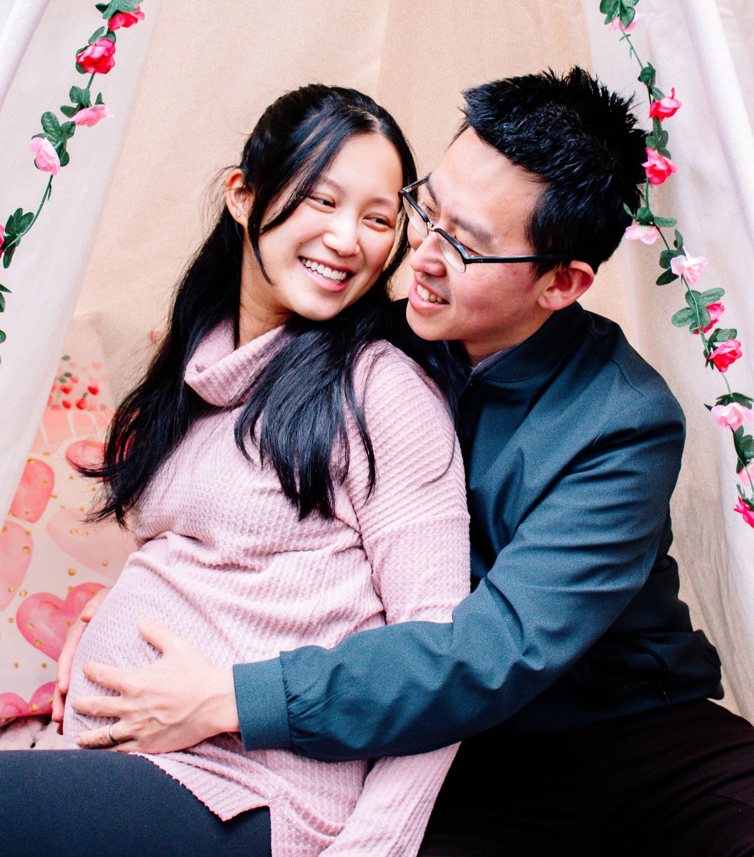 Expecting couple sharing a sweet moment under a floral teepee—joyful maternity mini session with soft pink tones. Maternity photography .