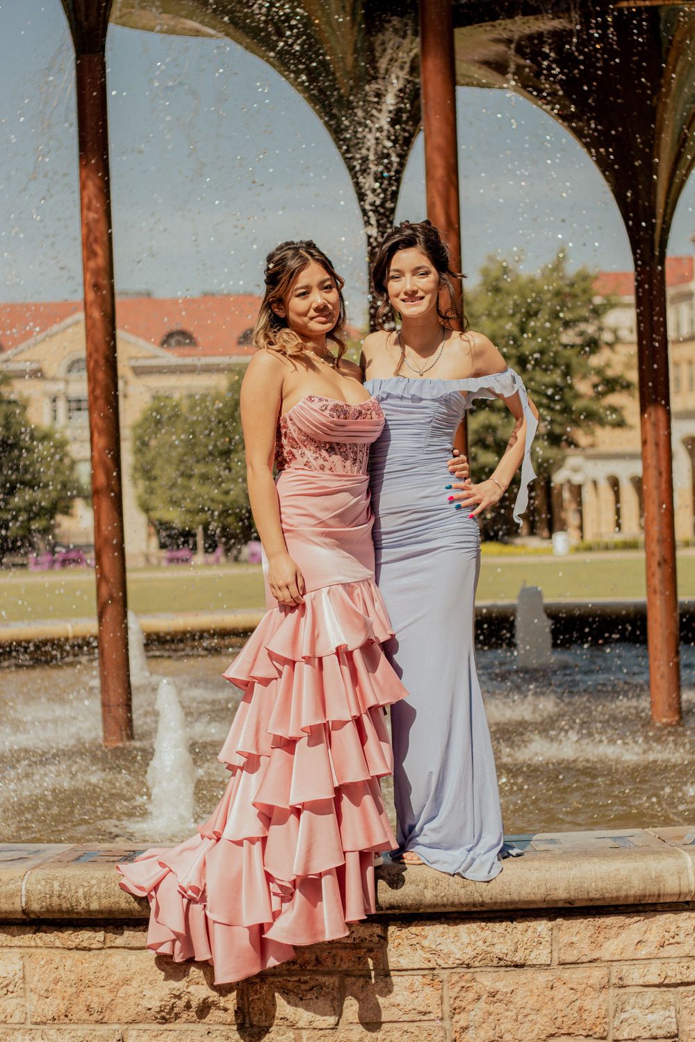 Two women in formal dresses, one pink with ruffles and one light blue, pose together by a fountain on a sunny day.