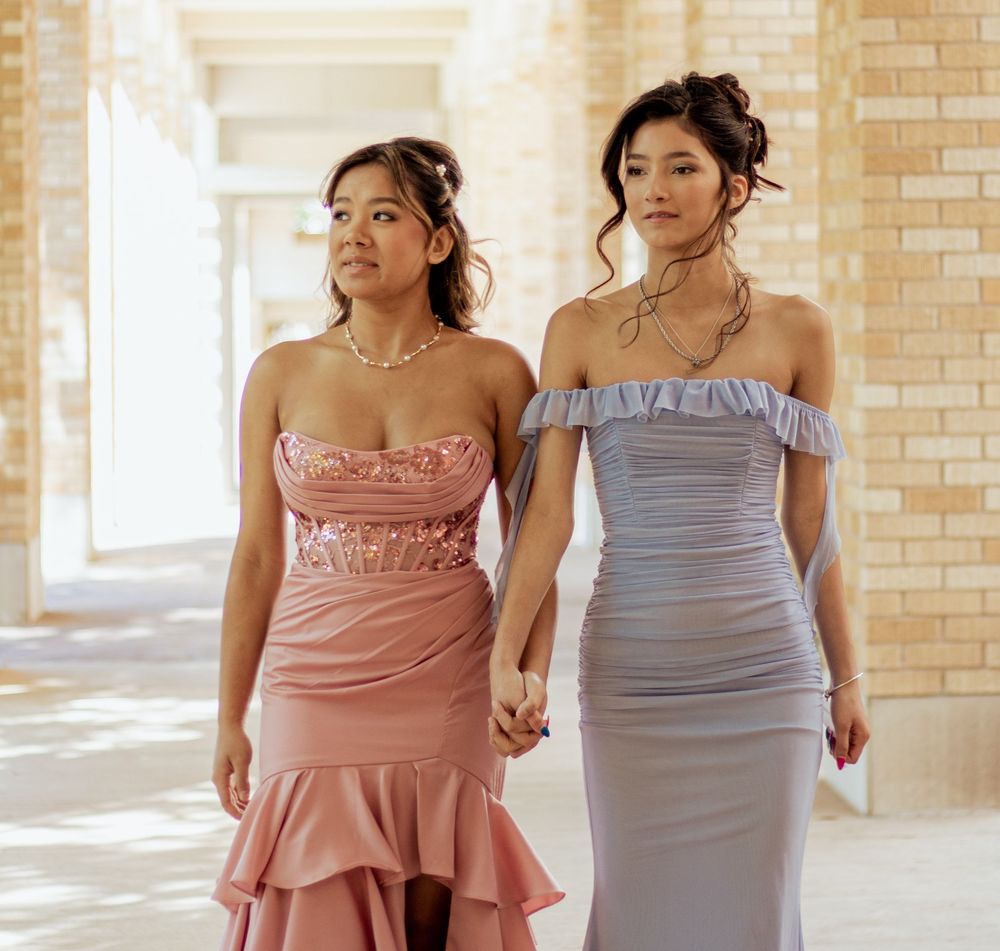 Two women in elegant evening gowns, one in blush pink with ruffles and one in light gray with off-shoulder detail.