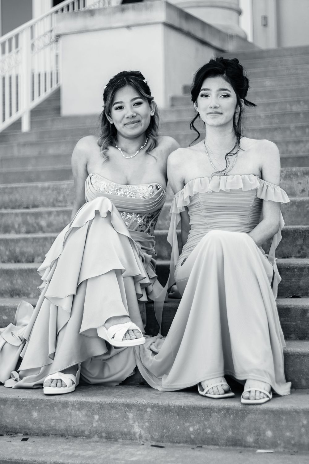 Two friends in elegant off-shoulder formal dresses sit together on stone steps in a black and white photograph.