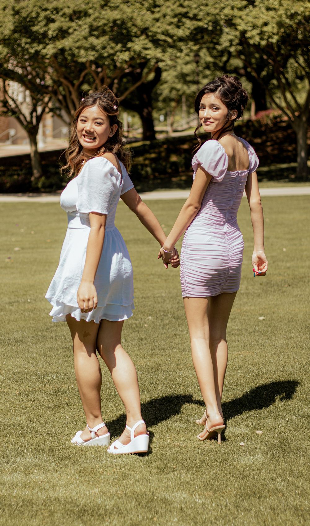 Two friends hold hands while walking on grass, one wearing a white dress and sneakers, the other in a pink fitted dress.