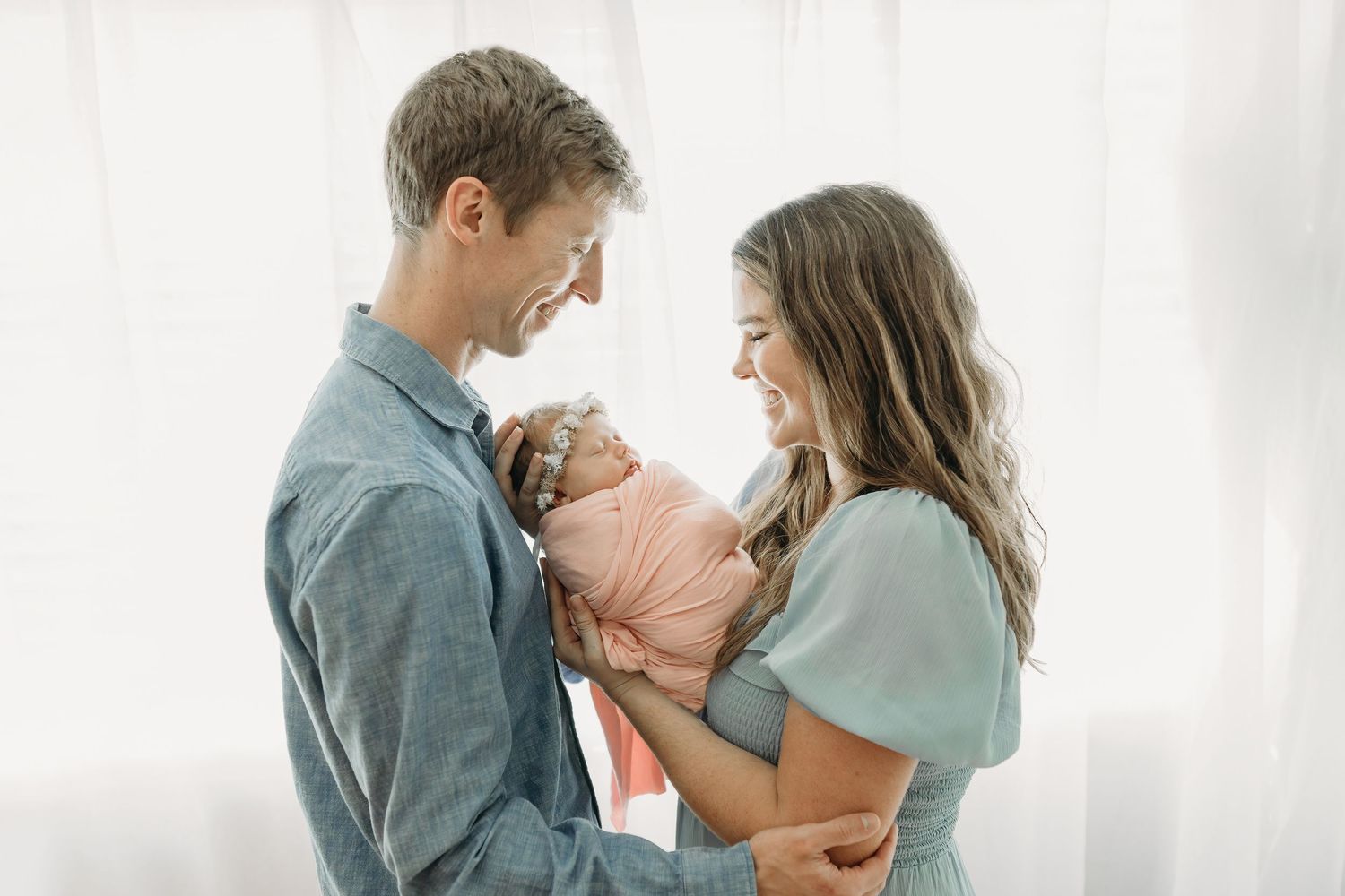 Parents wearing soft grey and mint colored clothing lovingly gaze down at their newborn baby against a white wall.