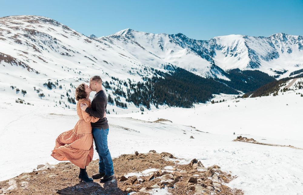 Couple embraces on snowy mountain overlook with dramatic Colorado Rocky Mountain peaks in background.