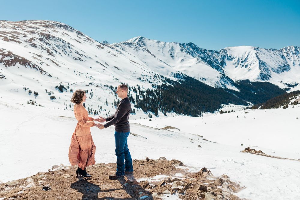 Two people stand together on a snowy mountain overlook with dramatic snow-capped peaks and blue sky in the background.