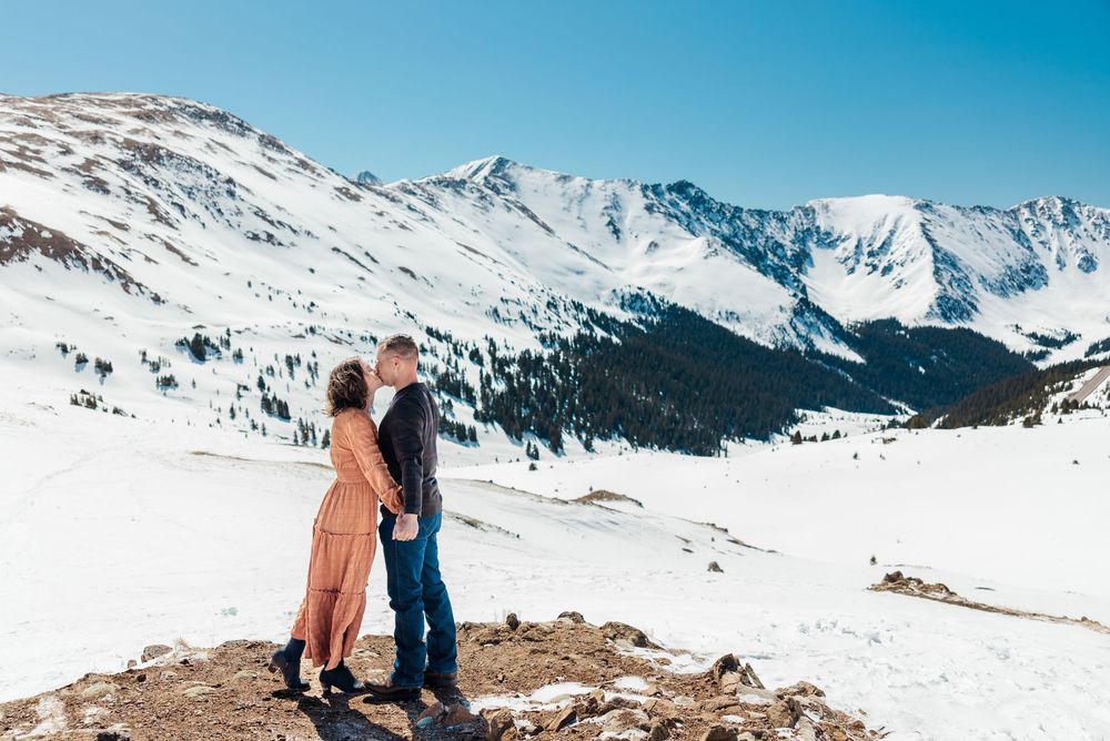 Two people stand on snowy mountain overlook with snow-capped peaks and blue sky in Colorado winter landscape.