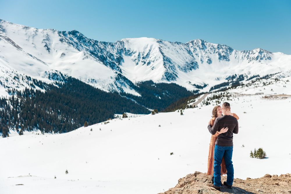 Snow-capped mountain peaks tower over a winter landscape with a couple standing on a scenic overlook.