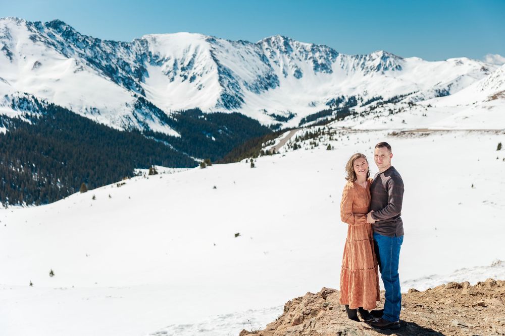 A couple stands together on a mountain viewpoint surrounded by snow-capped peaks and blue winter skies.