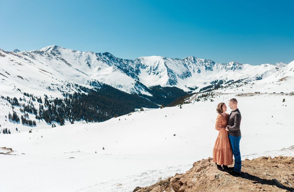 Two people embrace while overlooking a snow-covered mountain range and valley on a bright winter day in Colorado.