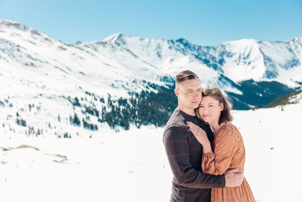A couple embraces in front of a snowy mountain landscape with white peaks and blue sky in the background.
