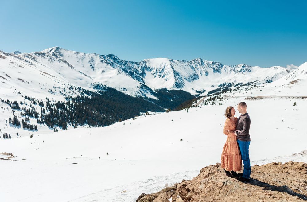 Couple stands on snowy mountain overlook with scenic snow-capped peaks and bright blue winter sky in background.