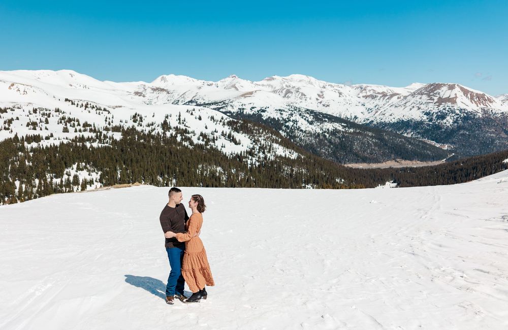 Two people in winter attire embrace while enjoying scenic snow-covered mountain views in a vast alpine landscape.