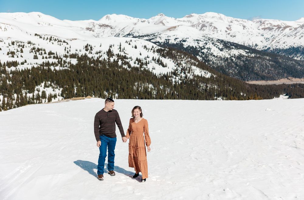 Two people walk together across a snowy mountain landscape with snow-capped peaks and evergreen forests in the distance.
