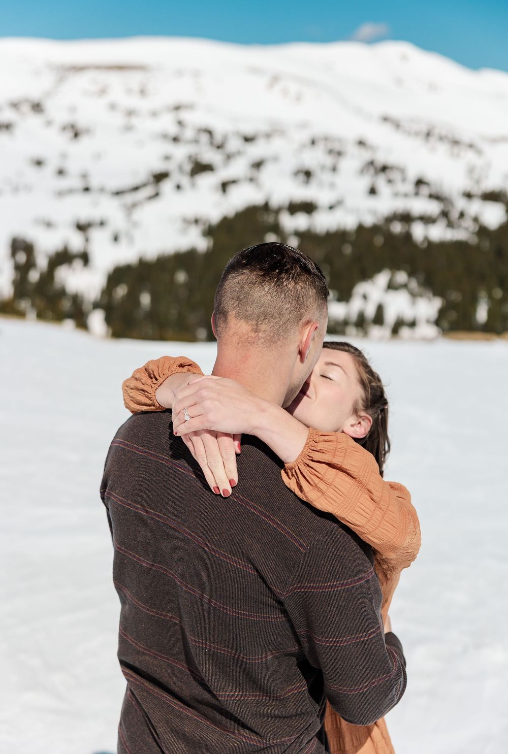 A romantic winter scene with a couple embracing in front of snow-covered mountains on a bright sunny day.
